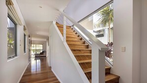Un escalier moderne avec des marches en bois et des rampes blanches, menant à une pièce lumineuse dotée de grandes fenêtres et offrant une vue sur des palmiers.
