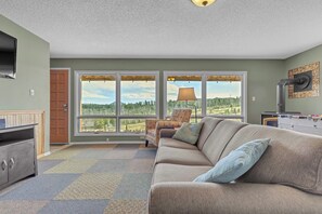 A living room with a sofa, a coffee table, a television, and a window with a view of a forest.