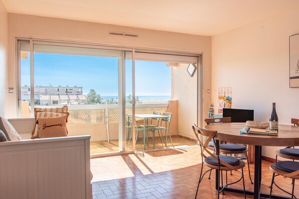 Un balcón con vista al mar, una zona de comedor con mesa y sillas, y un sofá con un cojín.