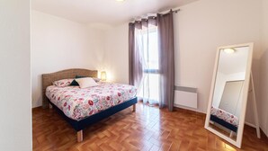 A bedroom with a bed, a bedside lamp, a window with curtains, a radiator, and a mirror. - The sound of the waves (Port-la-Nouvelle)