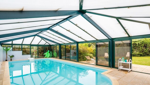 Una piscina cubierta con paredes de vidrio, un área de descanso con sillas y una mesa con objetos.
