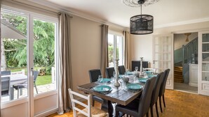 Comedor con mesa de madera dispuesta para seis personas, ventanas amplias y una escalera que sube a otro nivel.