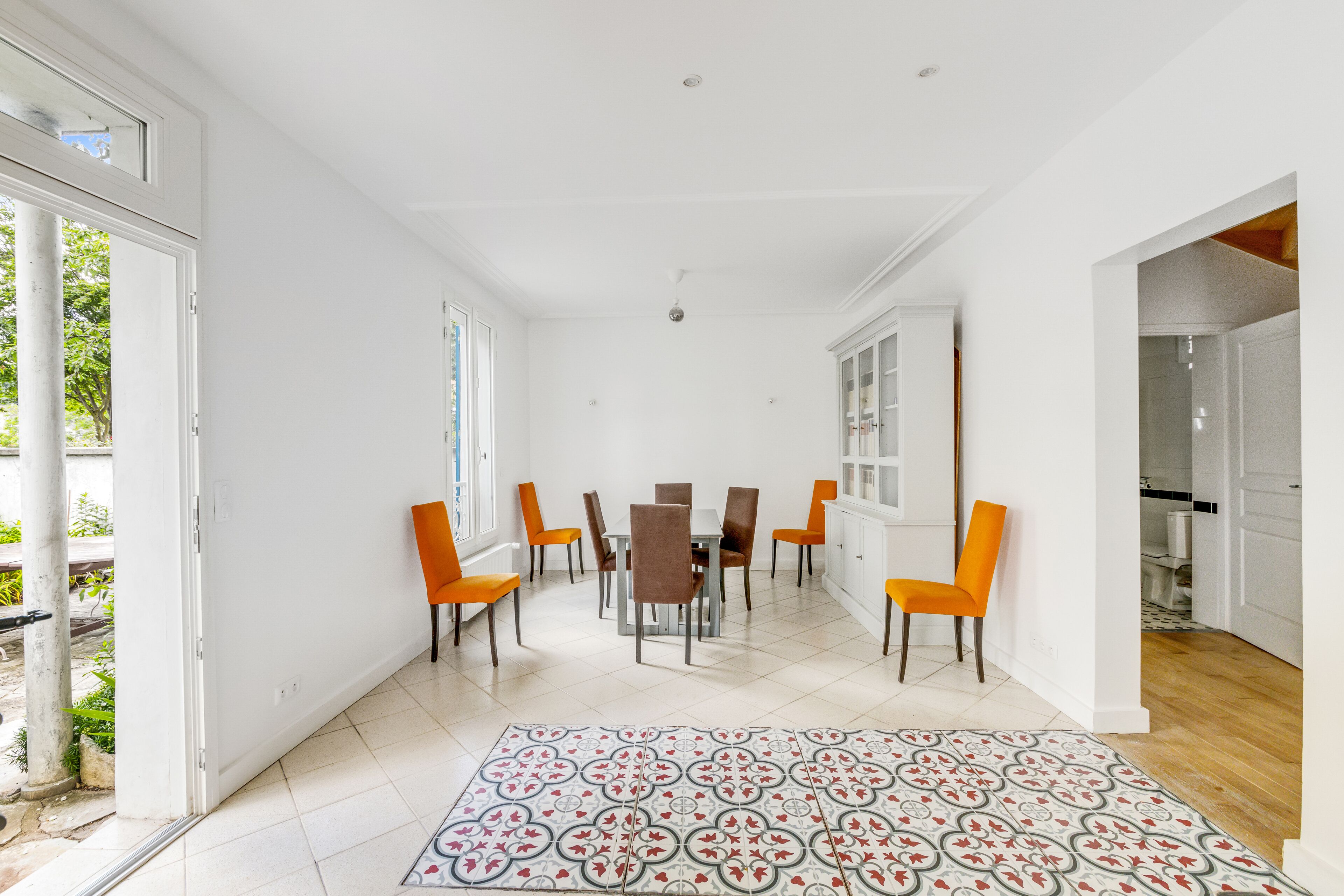 Een ruime eethoek met een witte betegelde vloer, een kamerkleed met dessin en oranje stoelen rondom een centrale tafel.