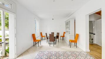Een ruime eethoek met een witte betegelde vloer, een kamerkleed met dessin en oranje stoelen rondom een centrale tafel.