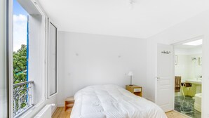 A bedroom with a large bed, a window with a view of greenery, a small bedside table with a lamp, and a bathroom visible through an open door.