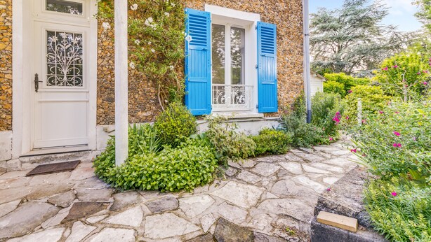 A stone house with a white door and blue shutters, surrounded by lush greenery and a stone pathway.