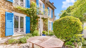 A stone building with blue shutters, a wooden table, and wicker chairs in a garden setting.