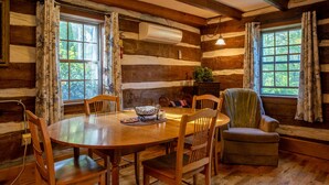 Interior rústico de una cabaña de madera con una mesa redonda de madera, cuatro sillas de madera, un sillón verde y dos ventanas con cortinas.