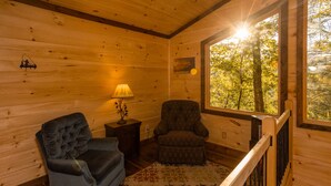 A cozy cabin interior with a wooden interior, a cushioned armchair, a smaller chair, a lamp, and a view of trees through a window. - Bufflehead | Cabin on the South Holston River (Bristol)