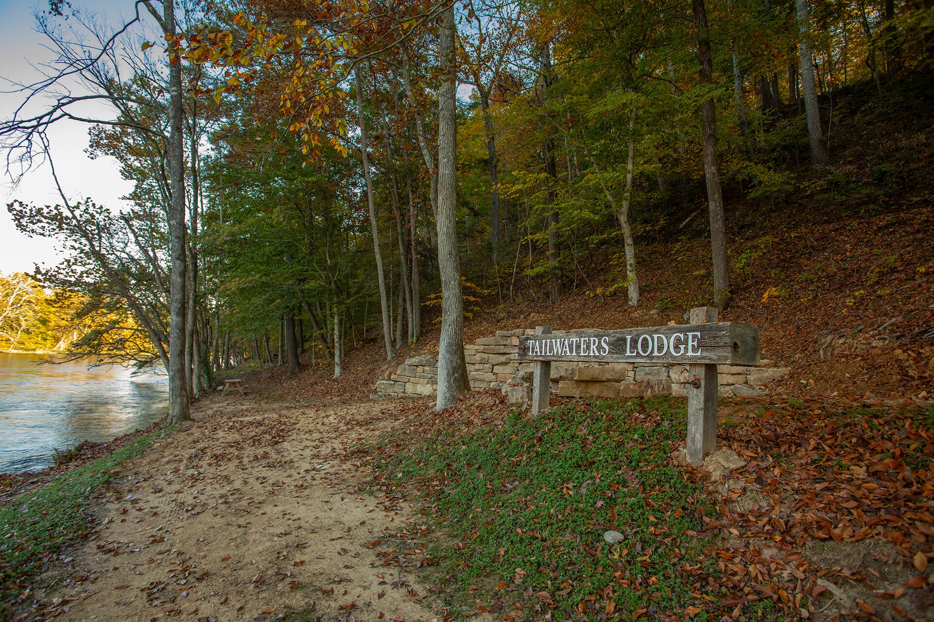 A wooden sign reading "Tailwaters Lodge" in a forested area with a dirt path leading to a body of water.