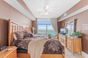 A bedroom with a bed, a desk, a ceiling fan, a window with a view of the ocean, and a mirror.