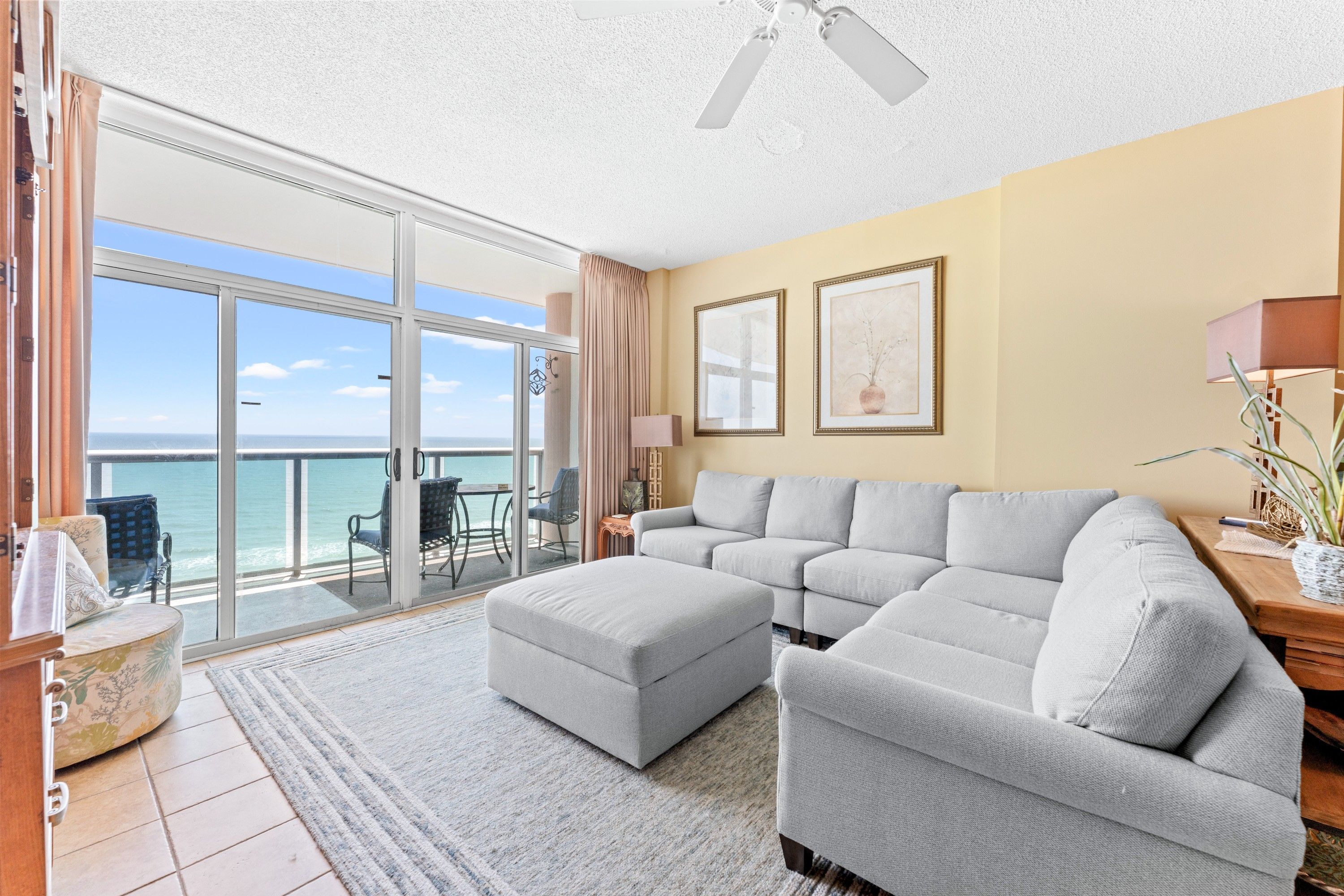 A modern living room with a large grey sectional sofa, a small ottoman, and a view of the ocean through a sliding glass door.