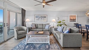 Un salon moderne avec des canapés gris, une table basse et un ventilateur de plafond. On y trouve deux lampes fixées au mur, un tableau encadré représentant une plage et une horloge au mur.