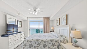 A bedroom with a bed, dresser, and nightstand, a ceiling fan, a window with a view, and artwork on the wall.