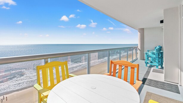 A balcony with a white table and chairs, overlooking a beach and the ocean.