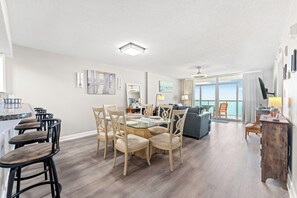 A spacious living area with a dining table, chairs, and a view of the sea through large windows.