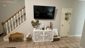 A living room with a wooden staircase, a flat-screen TV mounted on the wall, a white console table with decorative items, and a door with a floral wreath. - Townhome:  2 Bedrooms, 2.5 Baths, Close to Campus (Oxford)