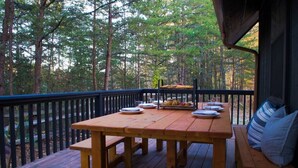 Terraza de madera con una mesa dispuesta para una comida, rodeada por una barandilla, con vista a un área boscosa.