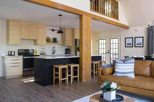 A modern kitchen with wooden cabinets, a central island with stools, and a dining area with a brown leather sofa.