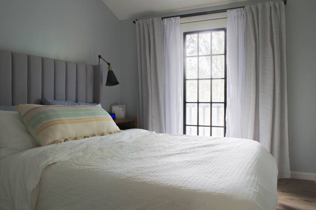 Una cama bien hecha con un almohadón a rayas, una lámpara de noche y una ventana con cortinas.