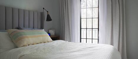 Una cama bien hecha con un almohadón a rayas, una lámpara de noche y una ventana con cortinas.