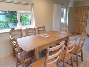 A dining area with a wooden table and chairs, a window with blinds, and a view of greenery outside.