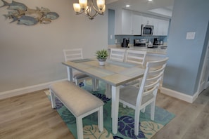 A dining area with a table, chairs, a bench, a chandelier, and a kitchen in the background.