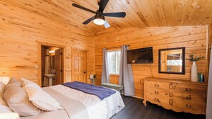 Ein Schlafzimmer in einer Holzhütte mit Bett, Kommode, Deckenventilator und einem Fenster mit Vorhängen.
