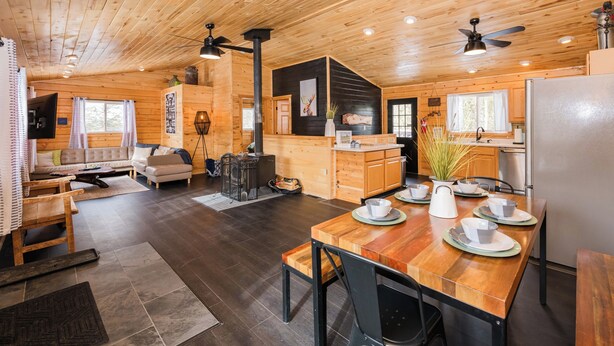 Ein Ferienhaus-Interieur mit einem Holzespeisetisch für vier Personen, einer Küchenzeile mit Kühlschrank und einem Wohnbereich mit Sofa und Kamin.
