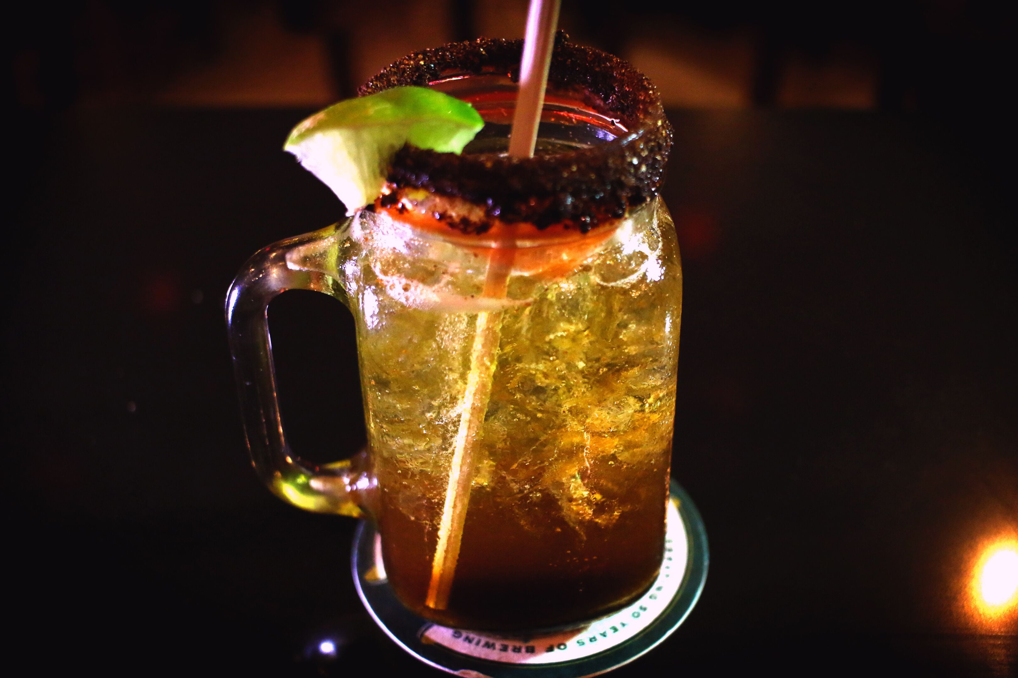Ein Glasbecher mit einer Limettenscheibe und einem salzigen Rand, gefüllt mit einer dunklen Flüssigkeit, möglicherweise ein Cocktail, auf einem Untersetzer mit Untersetzer-Logo.