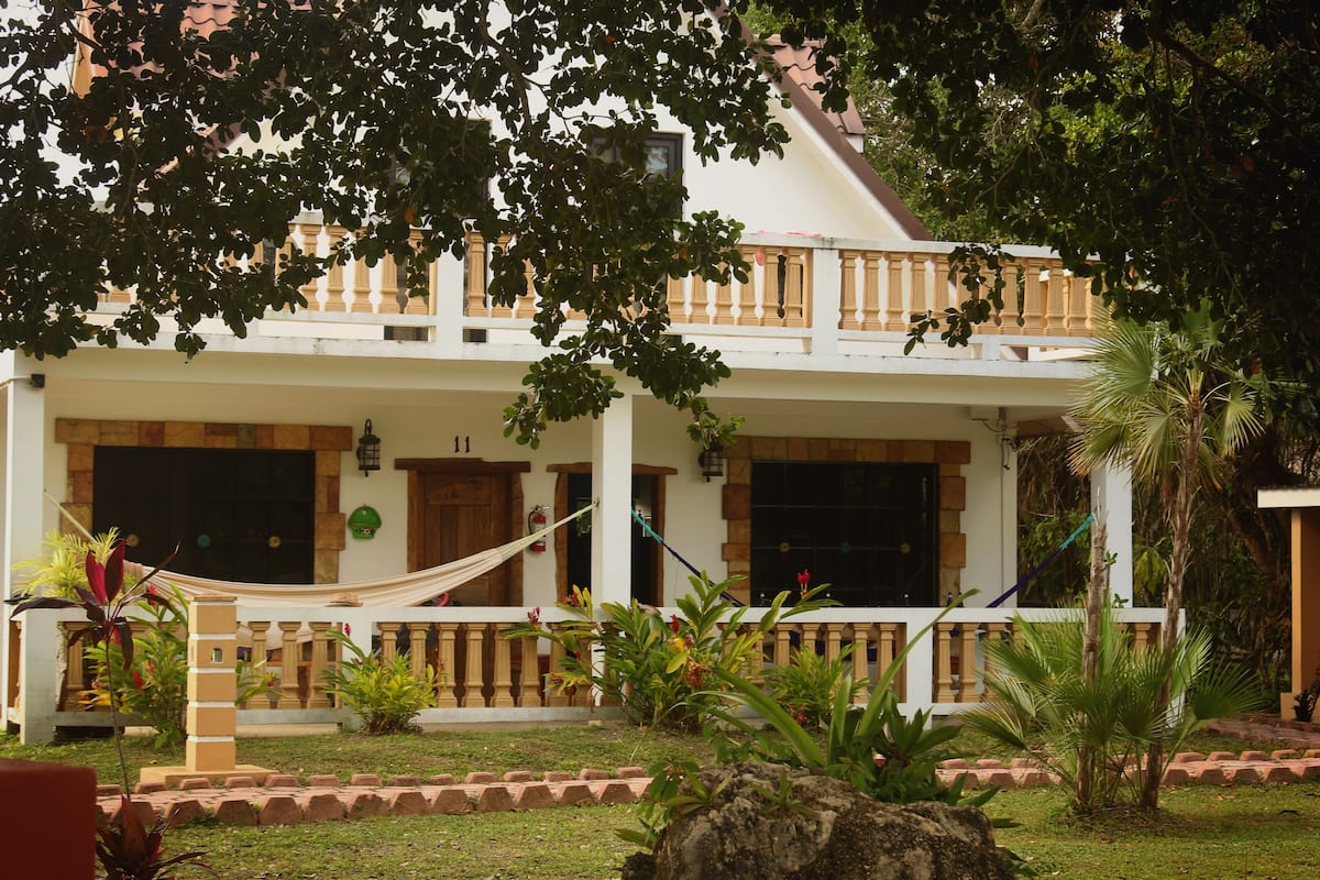 A two-story building with a balcony, surrounded by tropical plants and a hammock.