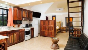 A kitchen with wooden cabinets, a stove, and a microwave. There is a black sofa and a wooden stool in the living area. - El Gran Mestizo Resort (Orange Walk)