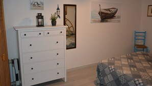 Une chambre avec une commode blanche, une couvre-lit à motifs, une chaise bleue et des œuvres d’art murales, notamment une peinture représentant un bateau et un oiseau.