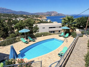 A swimming pool with lounge chairs and umbrellas, surrounded by stone tiles and a view of the sea and mountains.