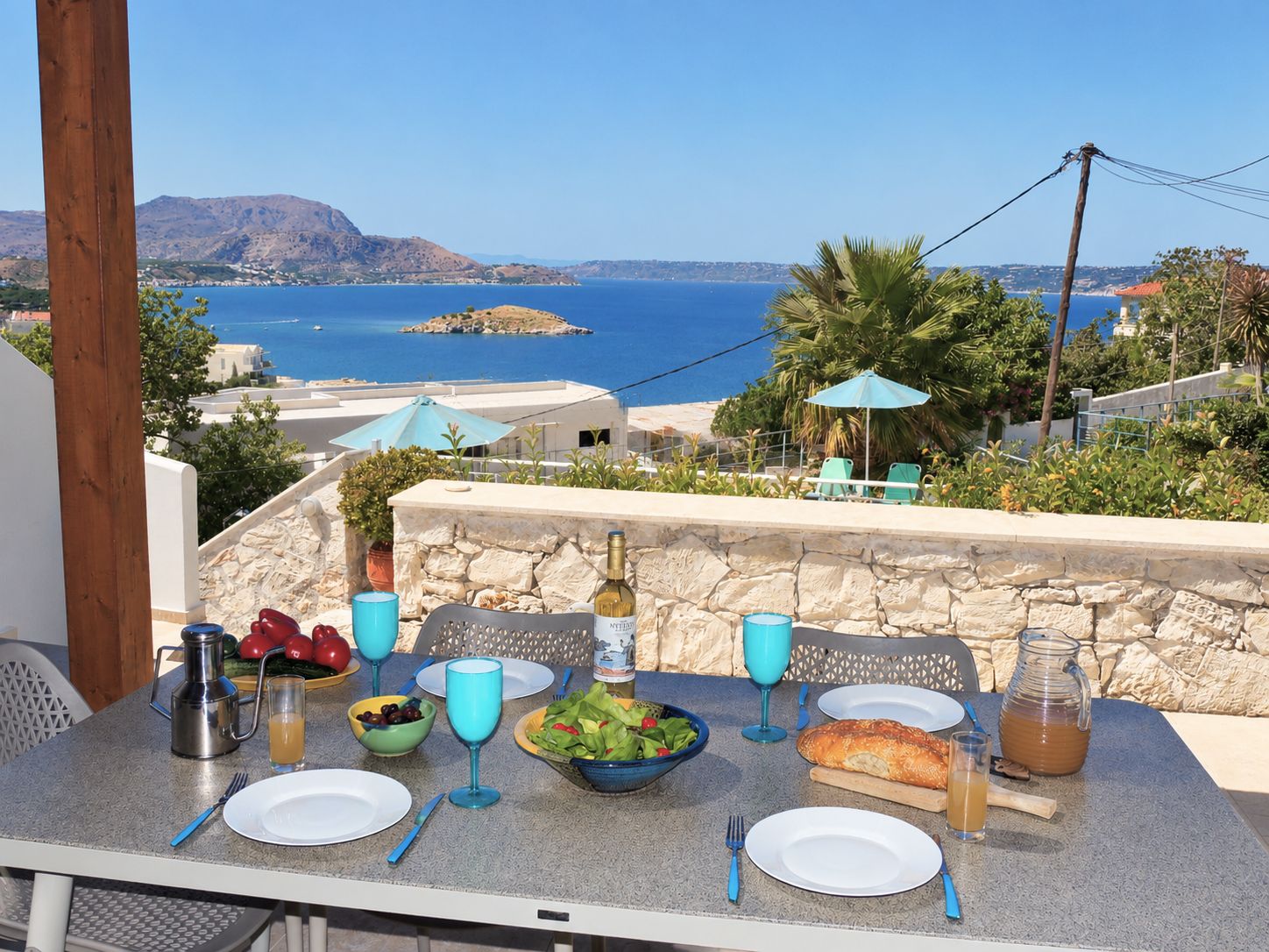 A table set with plates, glasses, and a bottle, overlooking a coastal view with mountains and a clear sky.