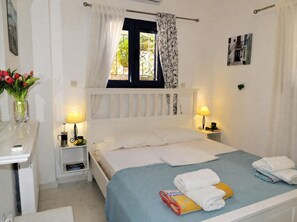 Une chambre à coucher comprenant un lit, deux tables de chevet, une fenêtre avec des rideaux, un tableau accroché au mur et un vase avec des fleurs.