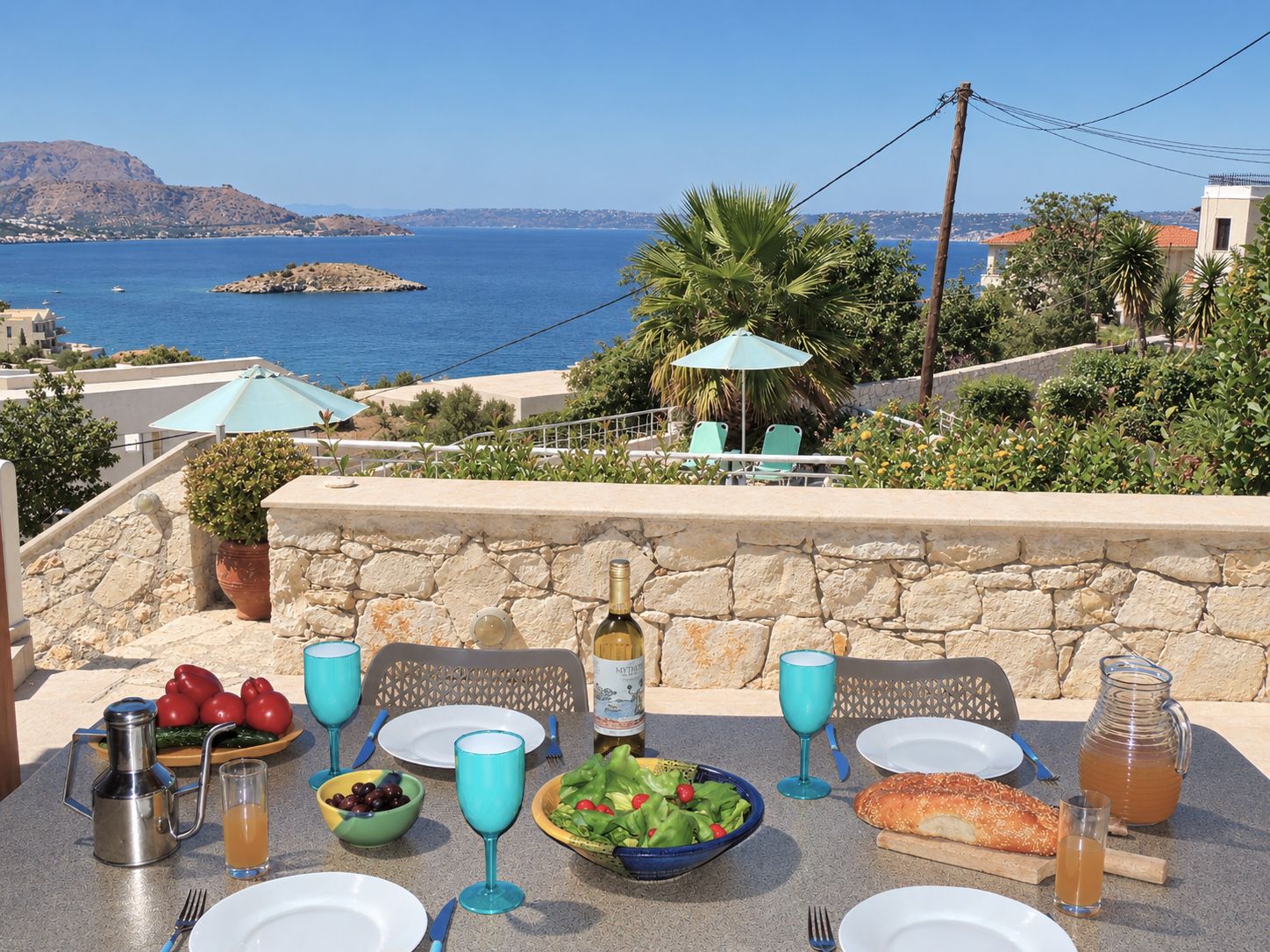 A table set with food and drinks, overlooking a coastal view with a stone wall and greenery.