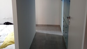 Een smalle gang met een betegelde vloer, een blauwe kast aan de rechterkant en een bed met een geel-witte deken in de linkerhoek.