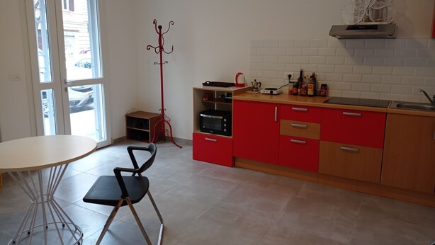 Een kleine keuken met een eethoek, met een witte tafel, een zwarte stoel, rode kastjes en een klein houten kastje.