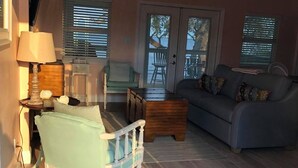 Una sala de estar acogedora con un sillón azul claro, un sofá gris y una mesa de centro de madera. Hay una lámpara sobre una mesita auxiliar y una puerta que da a un espacio exterior con una mesa y sillas.