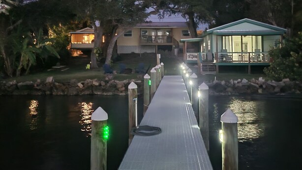 Un muelle de madera que lleva a una casa con porche cubierto, rodeada de árboles y un cuerpo de agua.