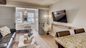 Una sala moderna con un sofá, una mesa de centro y una mesa de comedor. Hay un televisor montado en la pared y una puerta que da a otra habitación.