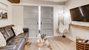 Una sala moderna con un sofá de cuero, una mesa de centro con objetos decorativos, un televisor montado en la pared y una puerta grande de color gris.