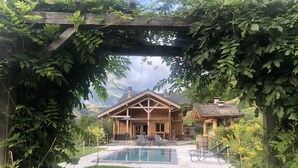 Una cabaña de madera con piscina, rodeada de exuberante vegetación y montañas al fondo.