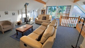 A spacious living room with a sofa, armchair, and a fireplace. There is a balcony with a railing and a view of greenery.