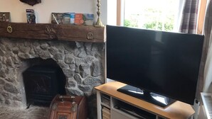 Un salon avec une cheminée en pierre, un téléviseur à écran plat posé sur un meuble en bois, un tapis rouge, une petite table ronde avec des livres et une horloge au mur.