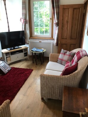 Een woonkamer met een televisie op een dressoir, een klein tafeltje met boeken, een rieten bank, een rood kleed en een houten deur.