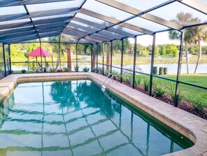 A swimming pool with a covered area, surrounded by a fence, and a view of a lake and greenery in the background. - Lake Front, Pool Home with Boat Dock (Inverness)