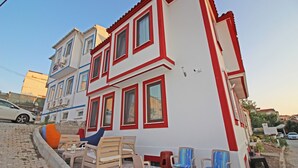 A two-story building with a red and white facade, outdoor seating area with multiple chairs, and a parked car in the background. - Narlı Pansiyon (Bozcaada)