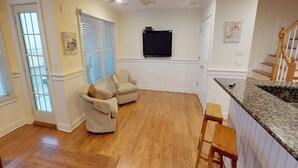 Een ruime woonkamer met houten vloer, een beige fauteuil, een houten salontafel en een keuken met een granieten aanrecht en barkrukken.